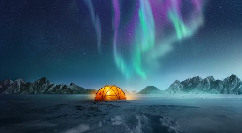 A tent pitched up in snow at night with the northern lights flickering in the sky above. Aurora Borealis and travelling. Photo composite.