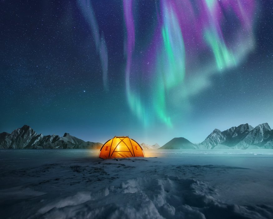 A tent pitched up in snow at night with the northern lights flickering in the sky above. Aurora Borealis and travelling. Photo composite.