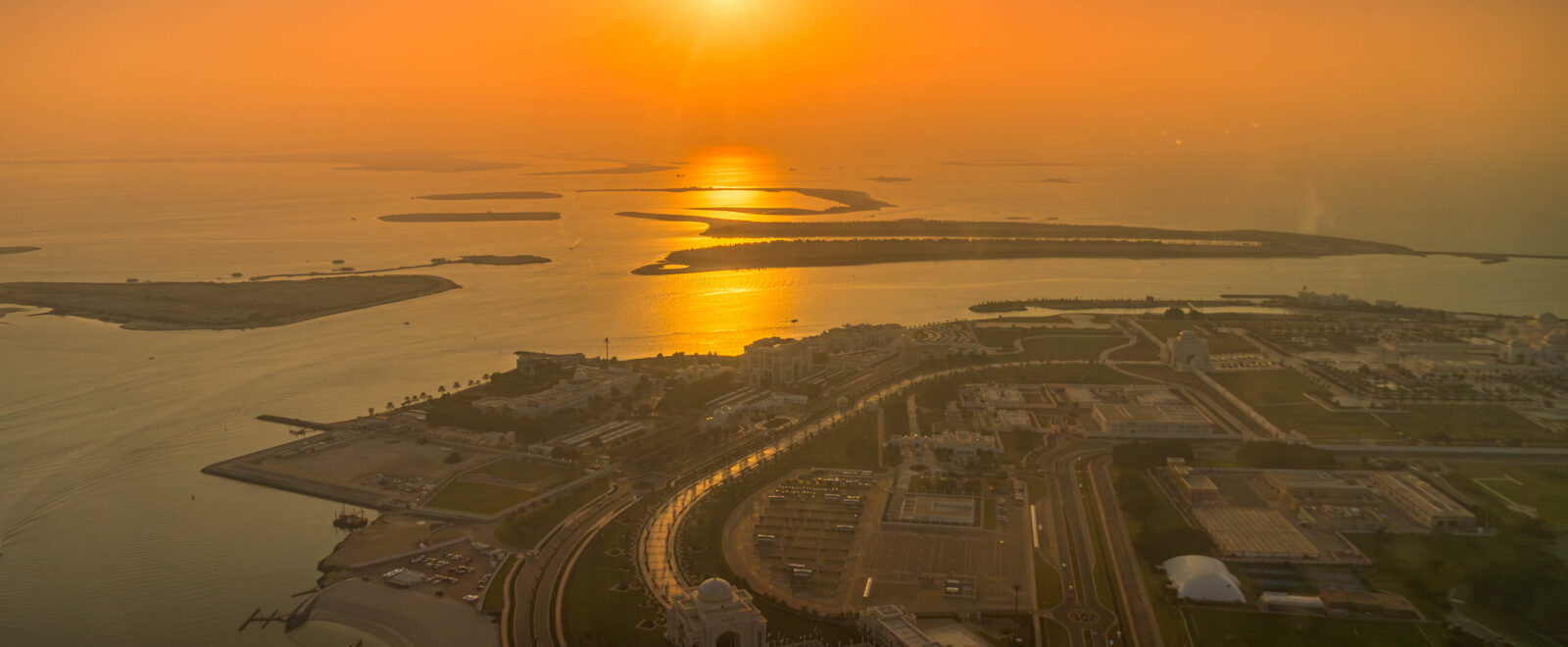 Aerial view of Abu Dhabi, UAE, including Qasr Al Watan and the Corniche. Coastal urban development meets the Persian Gulf during sunset glow.